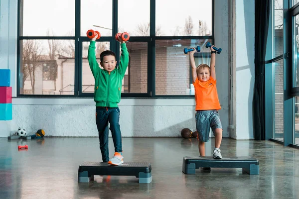 Vista frontal de niños multiétnicos levantando las manos con pesas mientras hacen aeróbicos Step en el gimnasio - foto de stock