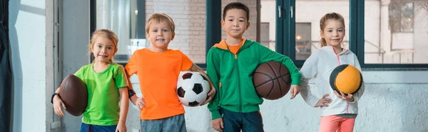 Vista frontal de niños multiculturales sosteniendo bolas y sonriendo juntos en el gimnasio, plano panorámico - foto de stock