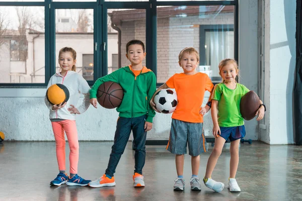 Vista frontale dei bambini multietnici che sorridono e tengono le palle in palestra — Foto stock