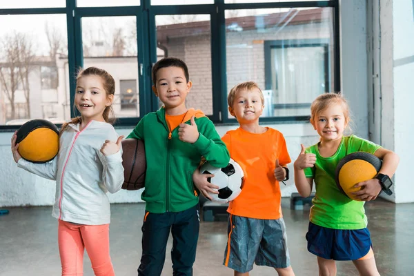 Vista frontal de niños felices multiculturales sosteniendo pelotas en el gimnasio - foto de stock