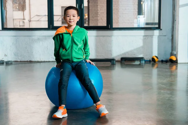 Vue de face de garçon asiatique assis sur la balle de fitness dans la salle de gym — Photo de stock