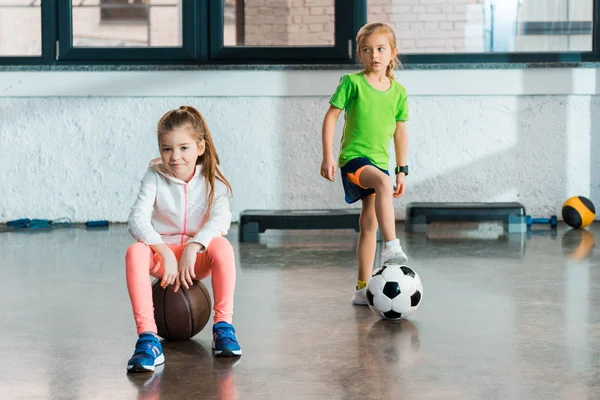 Frontansicht eines Kindes, das neben einem Kind auf einem Ball sitzt, das ein Bein auf einen Fußball setzt und in der Turnhalle wegschaut — Stockfoto