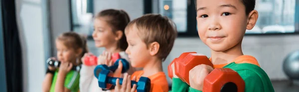 Concentration sélective des enfants multiethniques alignés, tenant des haltères dans la salle de gym, prise de vue panoramique — Photo de stock