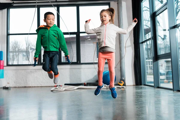 Vista frontal de niños multiculturales haciendo ejercicio con cuerda de salto en gimnasio - foto de stock