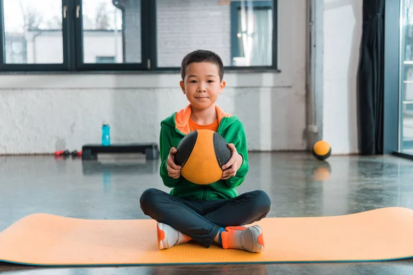 Selective focus of asian boy holding ball and sitting with crossed legs on fitness mat in gym — Stock Photo