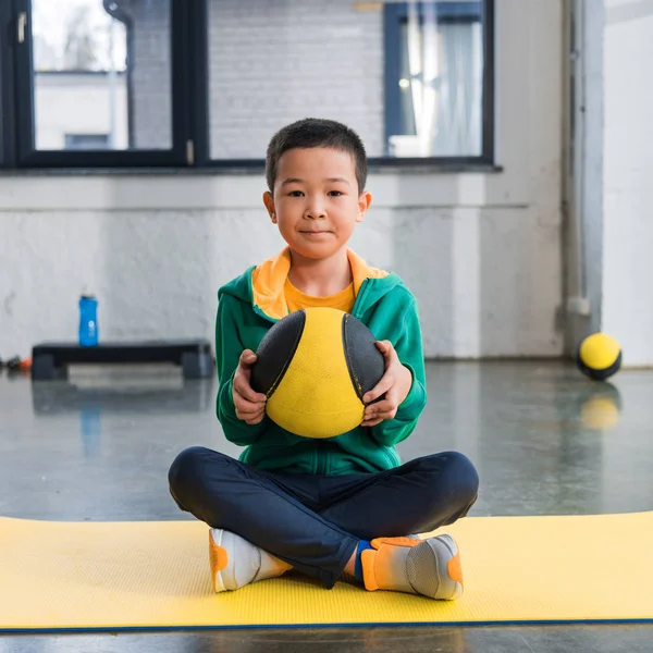 Selective focus of asian boy holding ball and sitting with crossed legs on fitness mat — Stock Photo