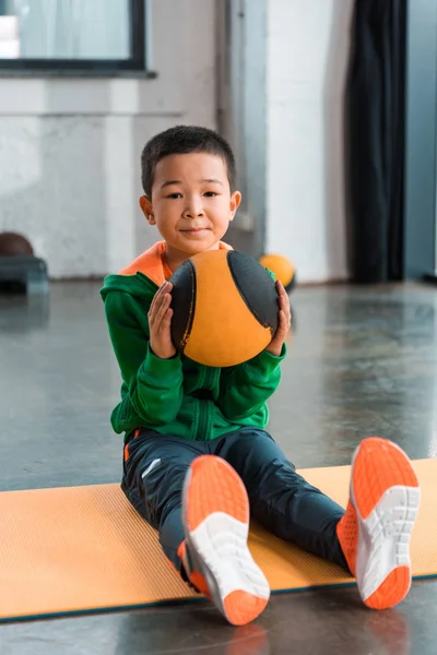 Селективный фокус азиатского мальчика, держащего мяч и сидящего на фитнес-коврике в спортзале — стоковое фото