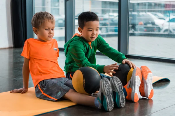 Enfants multiculturels regardant des balles sur les jambes et assis sur le tapis de fitness dans la salle de gym — Photo de stock