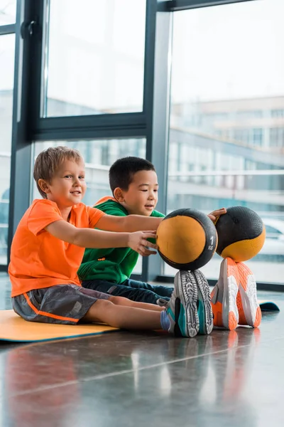 Niños multiculturales poniendo pelotas en las puntas de los dedos de los pies y sentados en la alfombra de fitness en el gimnasio - foto de stock