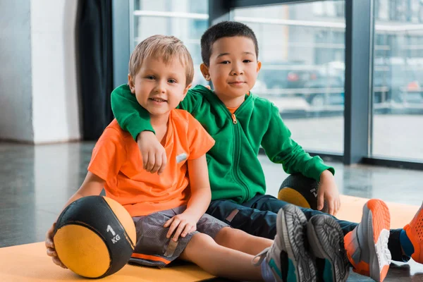 Multiethnic boys looking at camera, hugging and sitting with balls on fitness mat in gym — Stock Photo