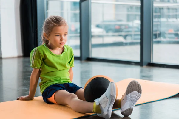 Kind mit Ball auf Beinen sitzt in Turnhalle auf Fitnessmatte — Stockfoto