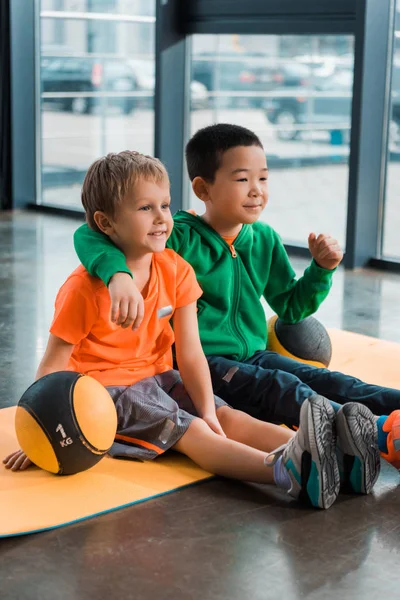 Multiethnic boys smiling, hugging and sitting with balls on fitness mat in gym — Stock Photo