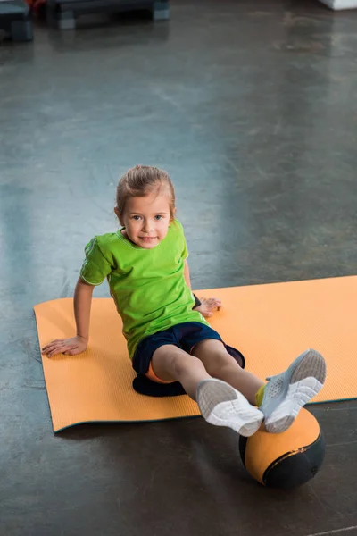 Criança com pernas na bola sentado no tapete de fitness no ginásio — Fotografia de Stock