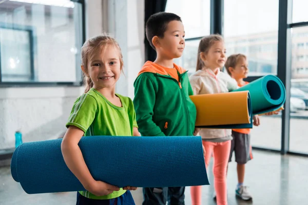 Foco seletivo de crianças multiculturais segurando tapetes de fitness no ginásio — Fotografia de Stock