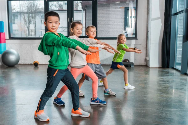 Vista laterale di bambini multietnici con le mani tese che fanno affondi in palestra — Foto stock