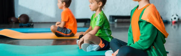Selective focus of children with crossed legs sitting on fitness mats in gym, panoramic shot — Stock Photo