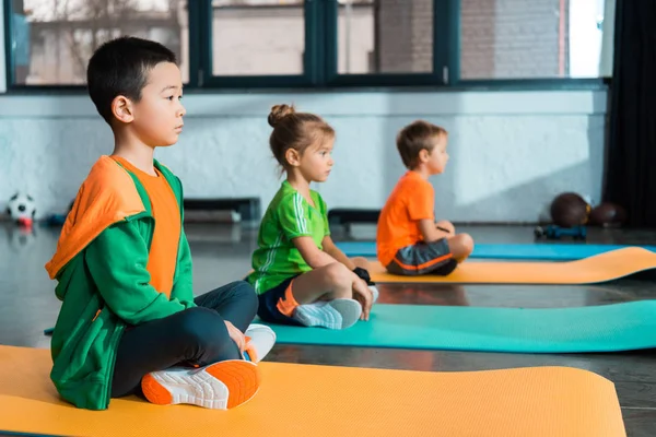 Selective focus of multicultural children with crossed legs sitting on fitness mats in sports center — Stock Photo