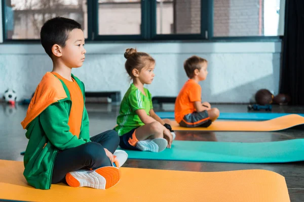 Enfoque selectivo de niños multiculturales con piernas cruzadas sentados en colchonetas de fitness en un centro deportivo - foto de stock