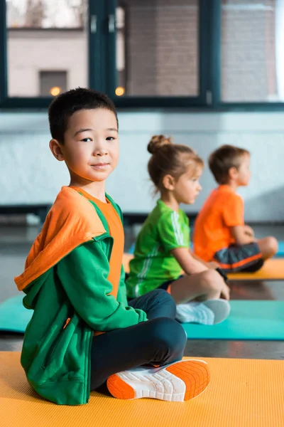 Focus selettivo di bambini multiculturali con gambe incrociate seduti su tappeti fitness in palestra — Foto stock