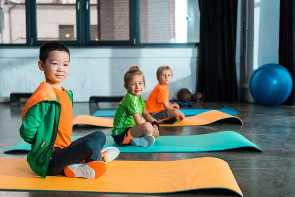 Enfoque selectivo de niños multiculturales con piernas cruzadas mirando a la cámara y sentados en colchonetas de fitness en el gimnasio - foto de stock