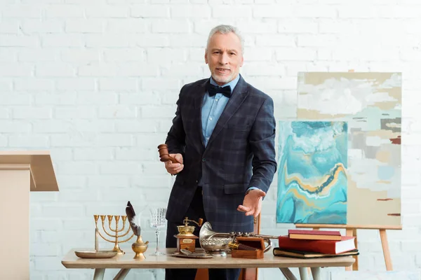 Encanteur souriant tenant un marteau et pointant à la main des objets anciens pendant la vente aux enchères — Photo de stock