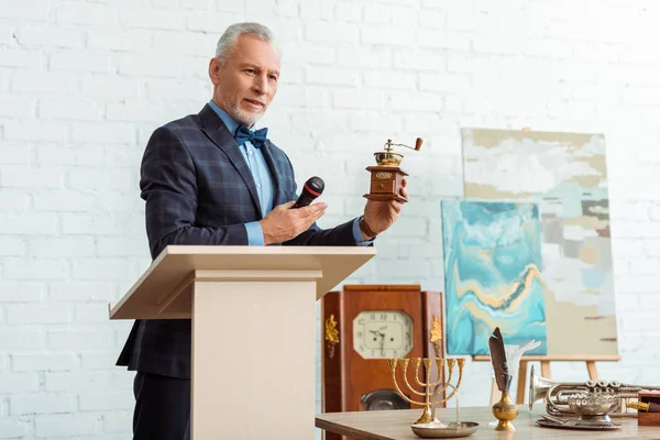 Belle encanteuse en costume pointant avec la main au moulin à café et tenant microphone pendant la vente aux enchères — Photo de stock