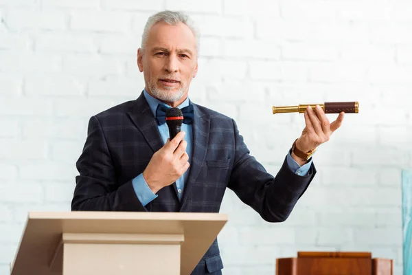 Handsome auctioneer in suit talking with microphone and holding spyglass during auction — Stock Photo