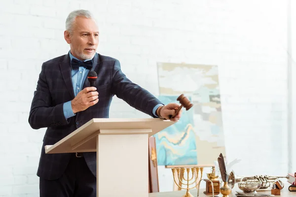 Belle encanteuse en costume parlant avec microphone et tenant gavel pendant la vente aux enchères — Photo de stock