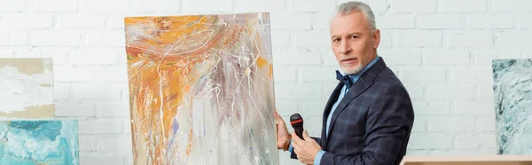 Panoramic shot of auctioneer talking with microphone and standing near picture during auction — Stock Photo