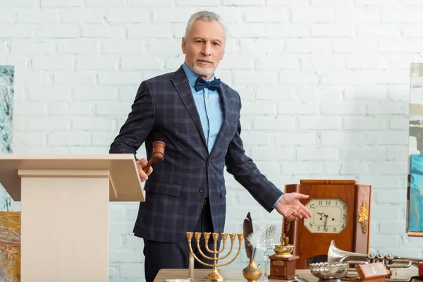 Handsome auctioneer holding gavel and pointing with hand at antique objects during auction — Stock Photo