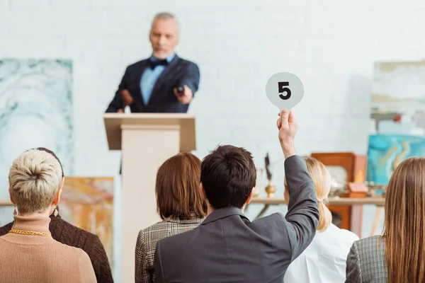 Back view of buyer showing auction paddle with number five to auctioneer during auction — Stock Photo