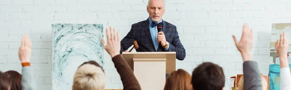 Panoramic shot of auctioneer holding gavel and microphone and looking at buyers with raised hands during auction — Stock Photo