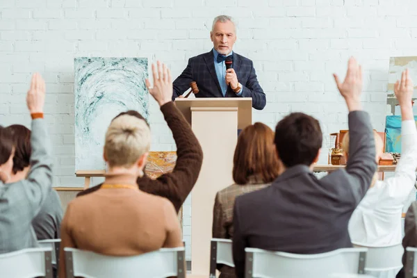 Selective focus of auctioneer holding gavel and microphone and looking at buyers with raised hands during auction — Stock Photo