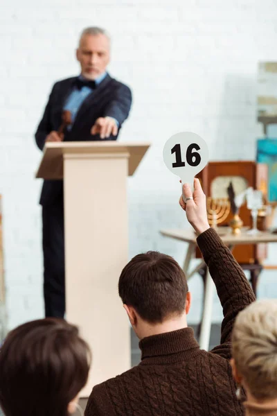 Back view of buyer showing auction paddle with number sixteen to auctioneer during auction — Stock Photo
