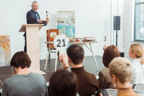 Selective focus of smiling auctioneer holding feather pen and pointing with finger at buyer during auction — Stock Photo