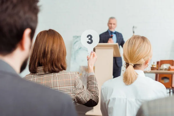 Back view of buyer showing auction paddle with number three to auctioneer during auction — Stock Photo