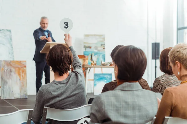 Back view of buyer showing auction paddle with number three to auctioneer during auction — Stock Photo