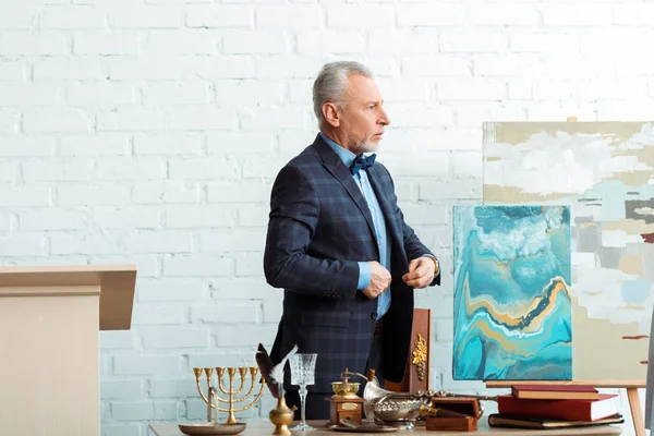 Handsome and mature auctioneer buttoning jacket near ancient objects — Stock Photo