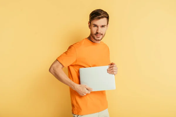 Handsome positive man looking at camera while holding closed laptop on yellow background — Stock Photo