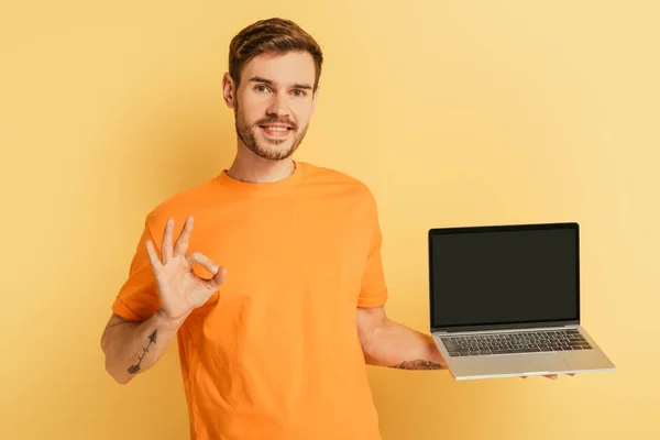 Усміхнений молодий чоловік демонструє добре жест, показуючи ноутбук з порожнім екраном на жовтому тлі — стокове фото