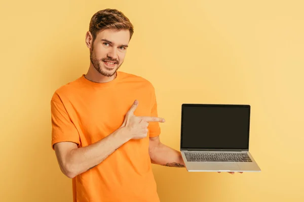 Joven alegre señalando con el dedo a la computadora portátil con pantalla en blanco sobre fondo amarillo - foto de stock