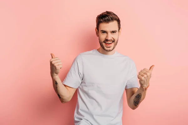 Glücklicher junger Mann zeigt Daumen nach oben, während er auf rosa Hintergrund in die Kamera lächelt — Stockfoto
