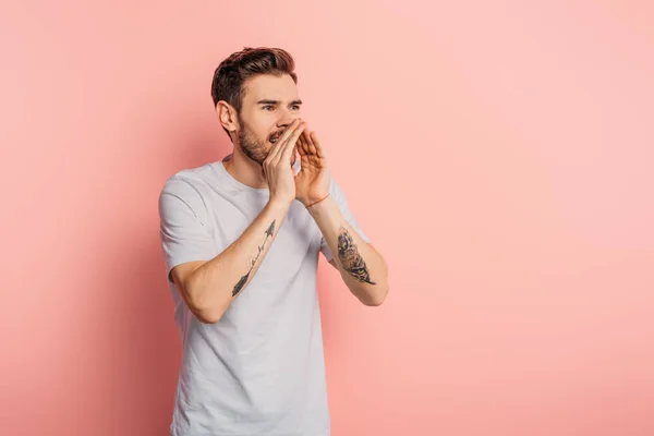 Excited young man screaming while holding hand near mouth on pink background — Stock Photo