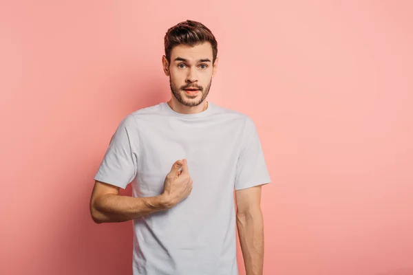 Sorpreso giovane uomo che punta con mano a se stesso mentre guarda la fotocamera su sfondo rosa — Foto stock