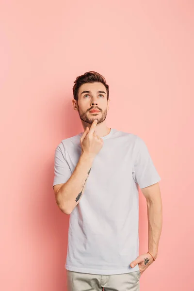 Pensive young man with hand in pocket touching chin and looking up on pink background — Stock Photo