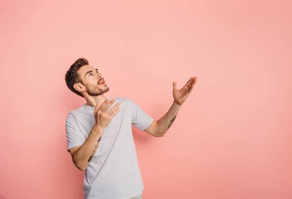 Amazed young man looking up while standing with open arms on pink background — Stock Photo