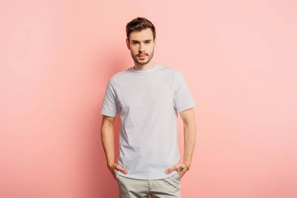 Joven guapo, seguro de sí mismo con las manos en los bolsillos mirando a la cámara en el fondo rosa - foto de stock