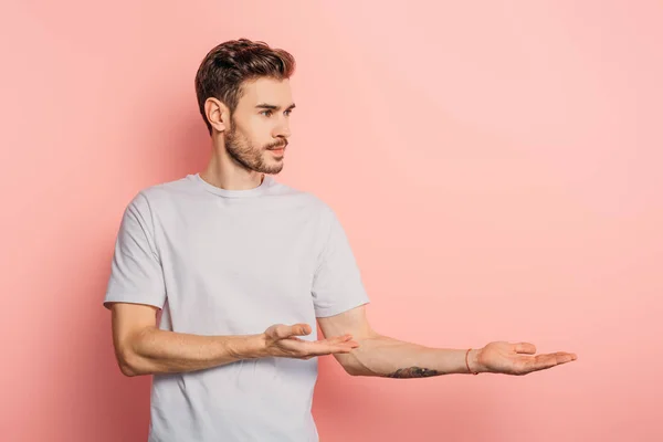 Schöner selbstbewusster Mann, der wegschaut und mit den Händen auf rosa Hintergrund zeigt — Stockfoto