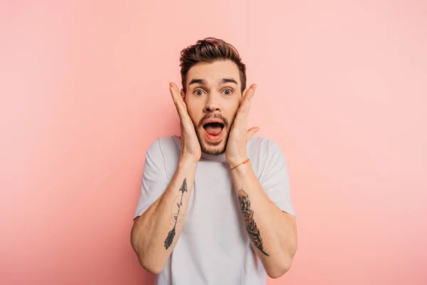 Shocked young man with open mouth and hands near face looking at camera on pink background — Stock Photo