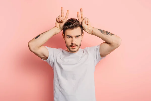 Ofendido joven mostrando cuernos gesto mientras mira a la cámara en el fondo rosa - foto de stock